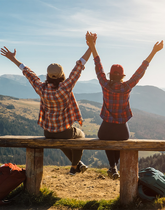 Couple on wooden mountain bench raising hands to the sky—active, healthy lifestyle